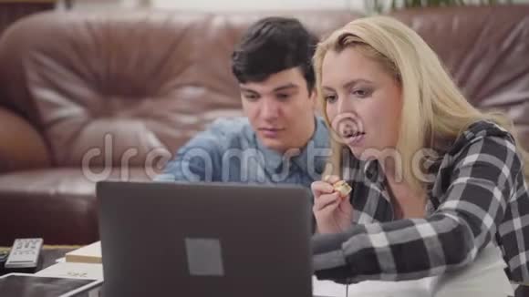 一对年轻夫妇坐在家里的笔记本电脑前一个黑发男人和一个金发女人一起在笔记本电脑前工作的肖像视频的预览图