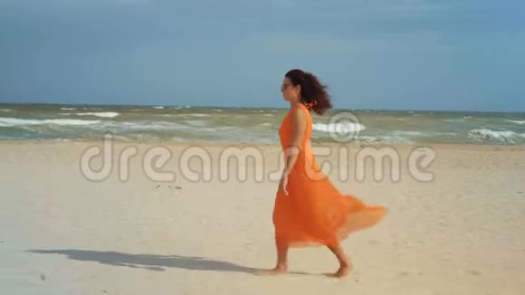穿着橘色连衣裙戴着太阳镜的美女赤脚踏上沙滩来到风和日丽的天气视频的预览图