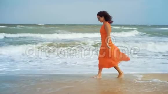 一位身穿橙色连衣裙戴着太阳镜的美女在风浪汹涌的大海背景下走在潮湿的沙滩上视频的预览图