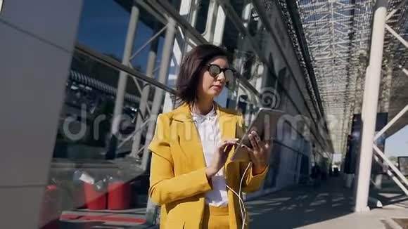 一位戴着耳机使用平板电脑的年轻漂亮女商人站在商业中心的城市街道上视频的预览图