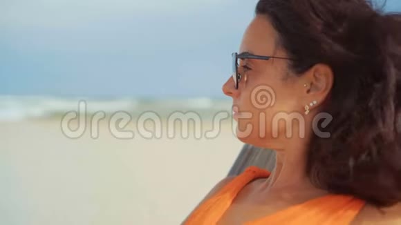 特写镜头中身着橙色连衣裙戴着墨镜的美女坐在沙滩上若有所思地向旁边看去视频的预览图