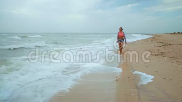 在暴风雨中赤脚漫步在海滩上的美丽女人手里拿着拖鞋视频的预览图
