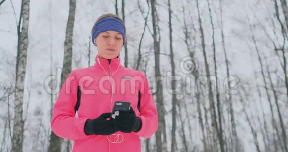 一位正在晨间慢跑的年轻女子手里拿着一部智能手机挑选一首音乐曲目进行训练冬季跑步使用视频的预览图