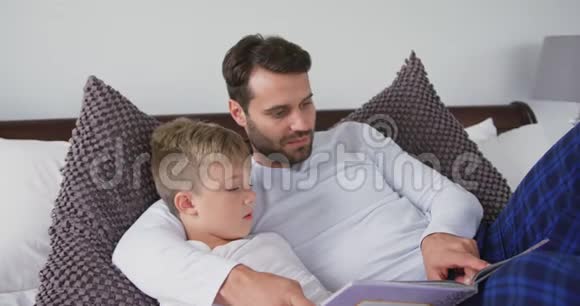 父亲和儿子躺在床上在家里的卧室里读一本故事书视频的预览图