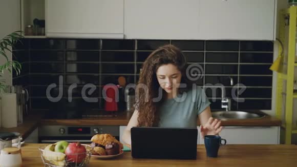 年轻迷人的卷曲快乐的白种人女人坐在厨房里用手提电脑喝茶她的丈夫来来来视频的预览图