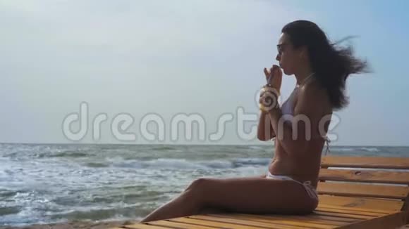 美丽的女人穿着白色泳衣和太阳镜坐在海边的木制躺椅上喝着一杯视频的预览图