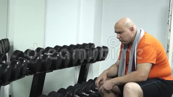 一个胖胖有趣的男人坐在健身房里无聊地看着哑铃视频的预览图