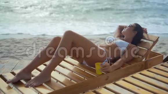 穿着白色泳衣戴着太阳眼镜的美女躺在海边的木制日光躺椅上视频的预览图
