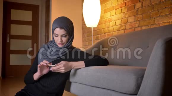 成年穆斯林女性戴着头巾坐在门口的地板上浏览电话时的特写照片视频的预览图