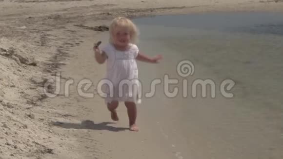 美丽的小女孩沿着海岸奔跑笑宝贝穿着白色连衣裙的小女孩在海边跑步慢动作视频的预览图