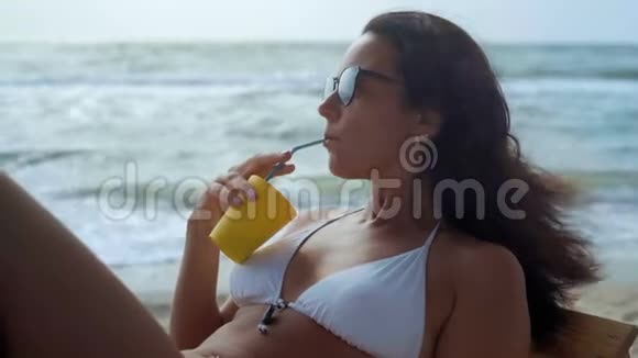 特写镜头一位身穿白色泳衣戴着太阳眼镜的美女躺在海边的木甲板椅上喝酒视频的预览图