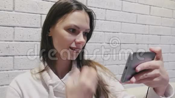 一位美丽的黑发女人在一堵砖白色的墙上看着一串戴着耳机的可怕的塞尔维亚语视频的预览图