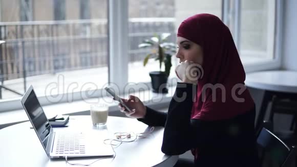 一个戴着传统阿拉伯头巾的可爱女孩正在她的笔记本电脑上做一些事情然后把耳机放进耳朵里视频的预览图