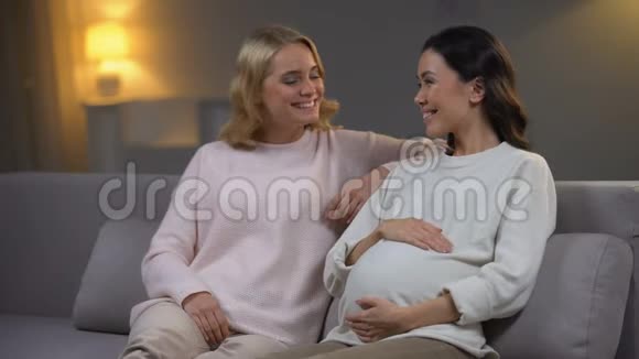年轻女子和怀孕的朋友坐在沙发上幸福的未来母亲视频的预览图