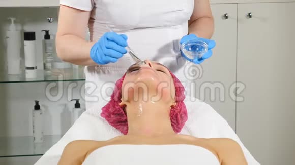 美容诊所年轻女人有专业的面部手术美容师用刷子在女性脸上涂面膜脸面视频的预览图