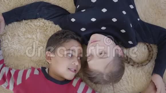 肖像可爱的非裔美国男孩和金发白种人女孩躺在米色蓬松的地毯上看着视频的预览图