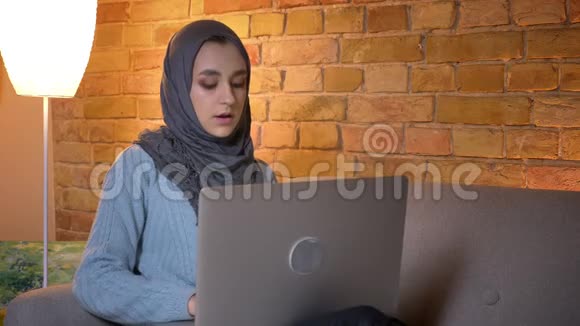 年轻迷人的穆斯林女性在舒适的室内沙发上坐着时使用笔记本电脑拍摄的特写镜头视频的预览图