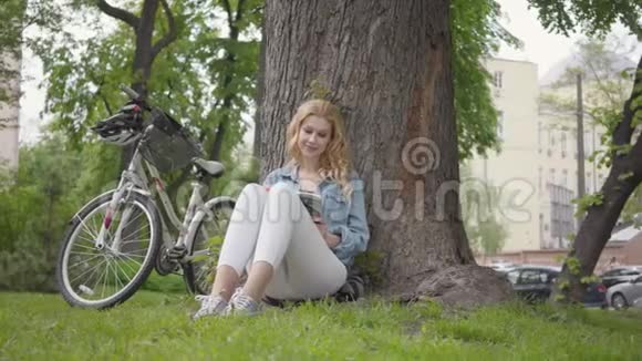 美丽的微笑的女人坐在公园的一棵老树下读日记现代自行车站在附近休闲视频的预览图