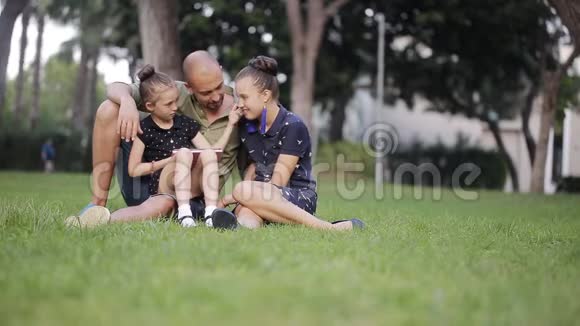 父亲带着两个女儿在草地上从平板电脑屏幕上读到故事笑了起来视频的预览图