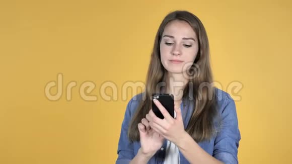 黄色背景下的漂亮女人应对损失和使用智能手机视频的预览图