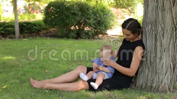 快乐的妈妈和她新生的儿子坐在一棵树下的夏天公园里视频的预览图