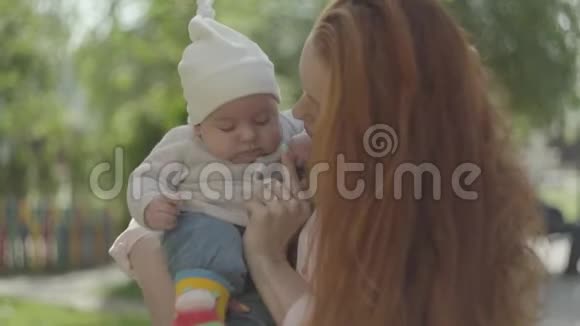 在春天的阳光明媚的日子里可爱的年轻漂亮的红头发妈妈抱着可爱的婴儿和他说话视频的预览图