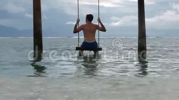 年轻的健康和迷人的男人坐在热带岛屿海滩度假胜地的海上秋千上望着地平线享受暑假视频的预览图