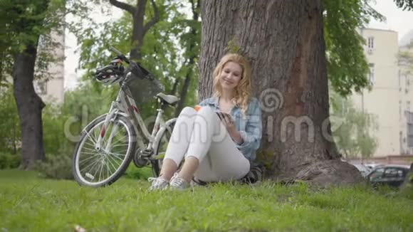 肖像可爱的微笑的女人坐在公园的一棵老树下读日记现代自行车站在附近视频的预览图