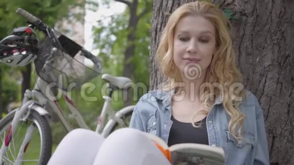 肖像可爱美丽年轻微笑的女人坐在公园的一棵老树下读日记现代自行车视频的预览图