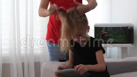 妈妈梳着可爱的女儿的头发她看着平板电脑妈妈和女儿在一起视频的预览图