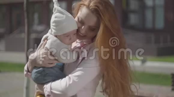 年轻漂亮的红发母亲抱着一个可爱的婴儿在春天阳光明媚的日子里和他聊天妈妈和视频的预览图