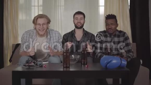 两个快乐的白种人和一个非裔美国人在家休息在电视上看拳击啤酒瓶空的视频的预览图
