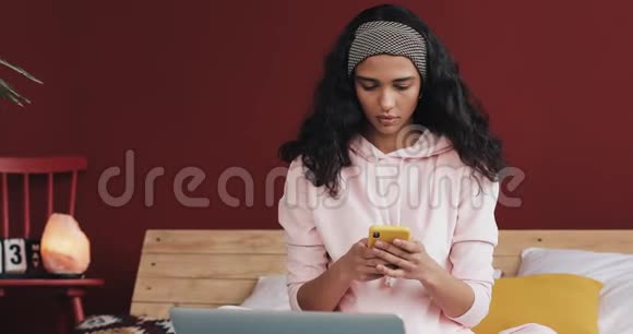穿着粉红色睡衣的漂亮年轻女子在家里坐在床上愤怒的嘴张着智能手机表达着情绪视频的预览图
