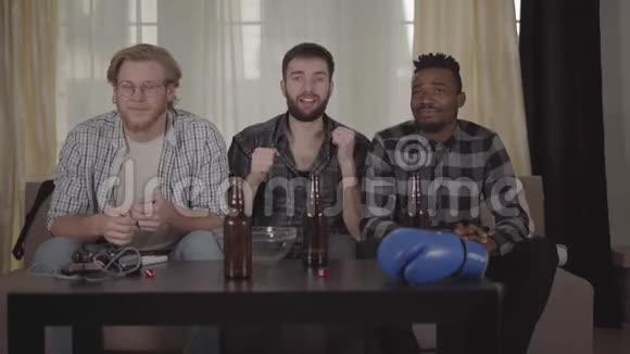 两个白种人和一个非裔美国人在家休息在电视上看拳击啤酒瓶空薯条视频的预览图