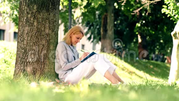 女孩坐在草地上的公园里在笔记本上做笔记学生正在公园里学习视频的预览图