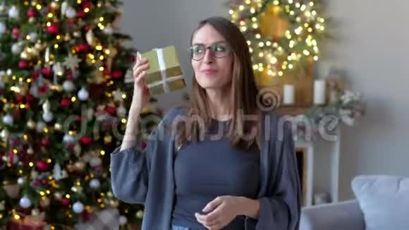 年轻兴奋的女人拿着礼物摇着盒子想猜猜里面是什么在圣诞树的背景上装饰着视频的预览图