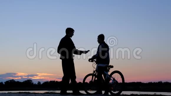 那家伙拿起电话在黎明时在电话上自拍一个男子和一个骑自行车的少年的剪影视频的预览图