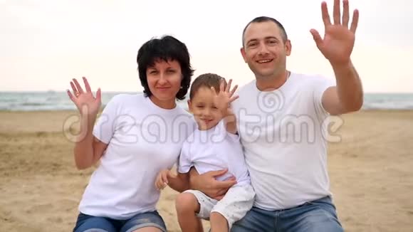 一个幸福的三口之家正坐在海边沙滩上微笑着向镜头挥手视频的预览图