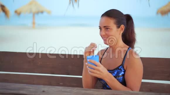在海边的一张木桌旁一位美丽的淡蓝色女人微笑着喝着鸡尾酒视频的预览图