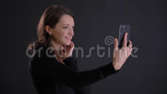 年轻迷人的高加索长发女孩在黑色背景下使用智能手机制作自拍照片的肖像视频的预览图