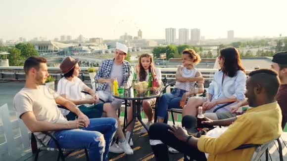 时髦的年轻人粗心的朋友坐在屋顶的桌子上开派对谈笑风生美丽的城市视频的预览图
