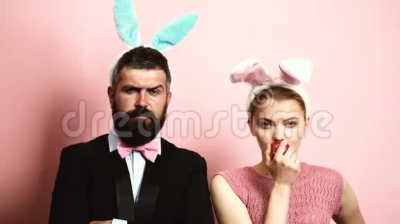 有胡子的男人有耳朵兔子和金发女人有耳朵兔子在粉红色的背景上吃苹果有兔子耳朵的女人视频的预览图