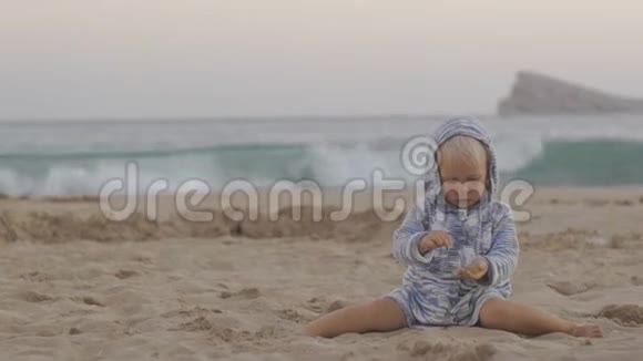穿着针织连帽衫的小女孩在暴风雨的海上背景下的日落海滩上玩沙子视频的预览图