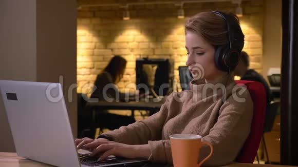 年轻的短发女士戴着耳机在办公室里专心地在笔记本电脑上打字和喝饮料视频的预览图