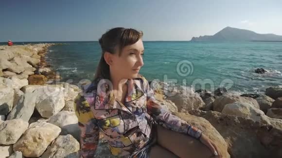 年轻迷人的女人微笑着坐在岩石上的夕阳山和海岸背景视频的预览图
