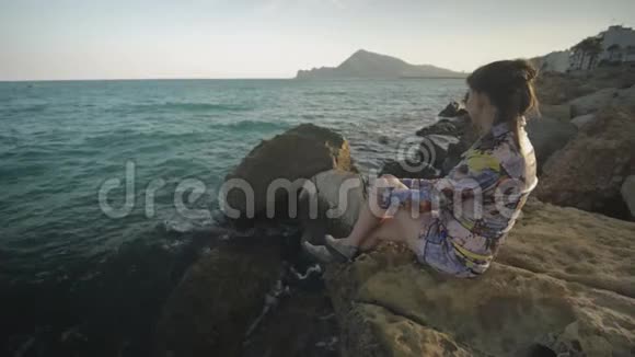 年轻迷人的女人坐在石头上眺望远处的夕阳山和海岸背景视频的预览图