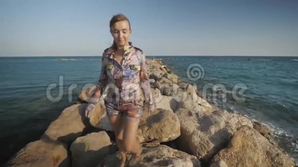 年轻迷人的女人在海边的石头上行走头发随风飘动视频的预览图