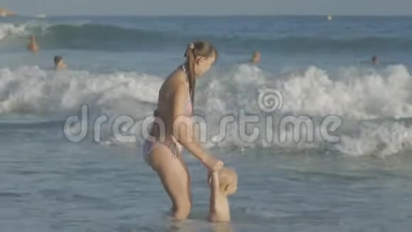 年轻的白种人母亲和两岁的金发碧眼的婴儿在海边玩水浪溅的游戏视频的预览图