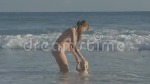 年轻的白种人母亲和两岁的金发婴儿一起玩耍在海浪上跳跃手牵着手在海边快乐视频的预览图