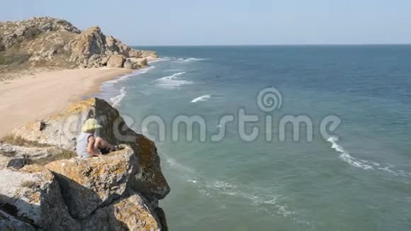 男孩坐在悬崖边上波浪冲刷沙质海岸山脊在岩石上破碎了视频的预览图
